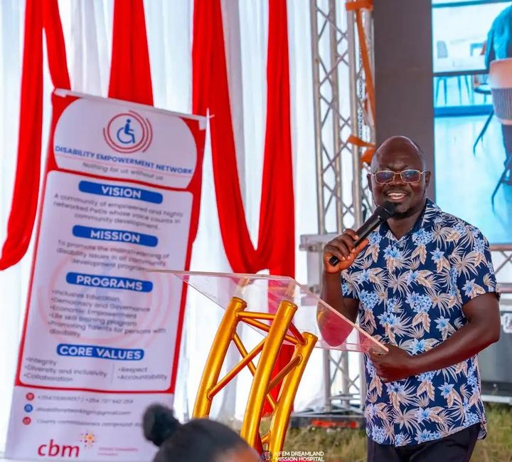 DEN CEO Adrian Sakwa smiling as he speaks into a microphone at the Phase 2 project launch, beside a DEN banner showing its vision, mission, programmes and core values