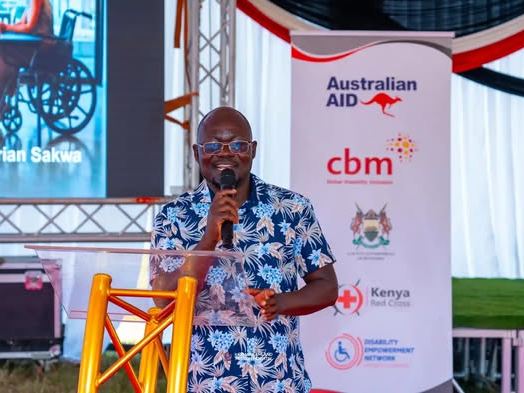 Adrian Sakwa, DEN CEO, speaking into a microphone at the podium beside partner banners