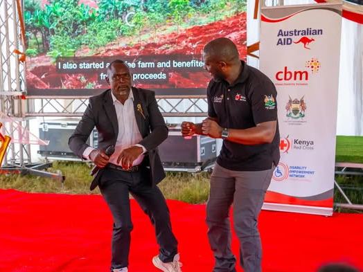 Two men on stage at the launch, one holding a microphone, in front of a slide about a farm started from project proceeds